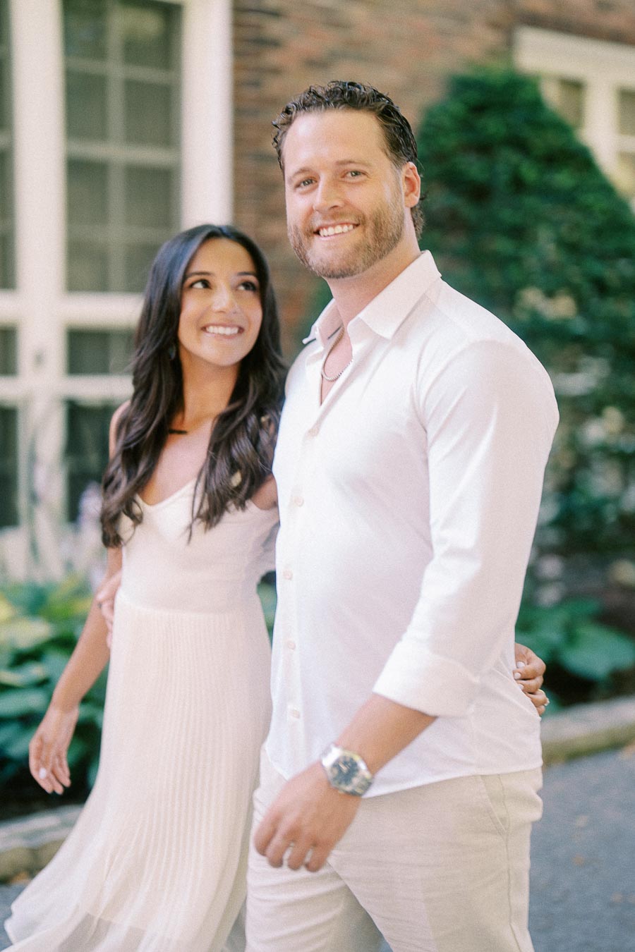 Rachel smiles up at Matt as they walk together beside a brick building and garden greenery.