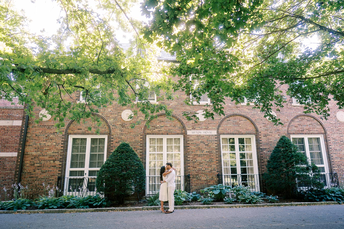 Rachel and Matt embrace beneath leafy trees in front of a brick campus building with tall windows.