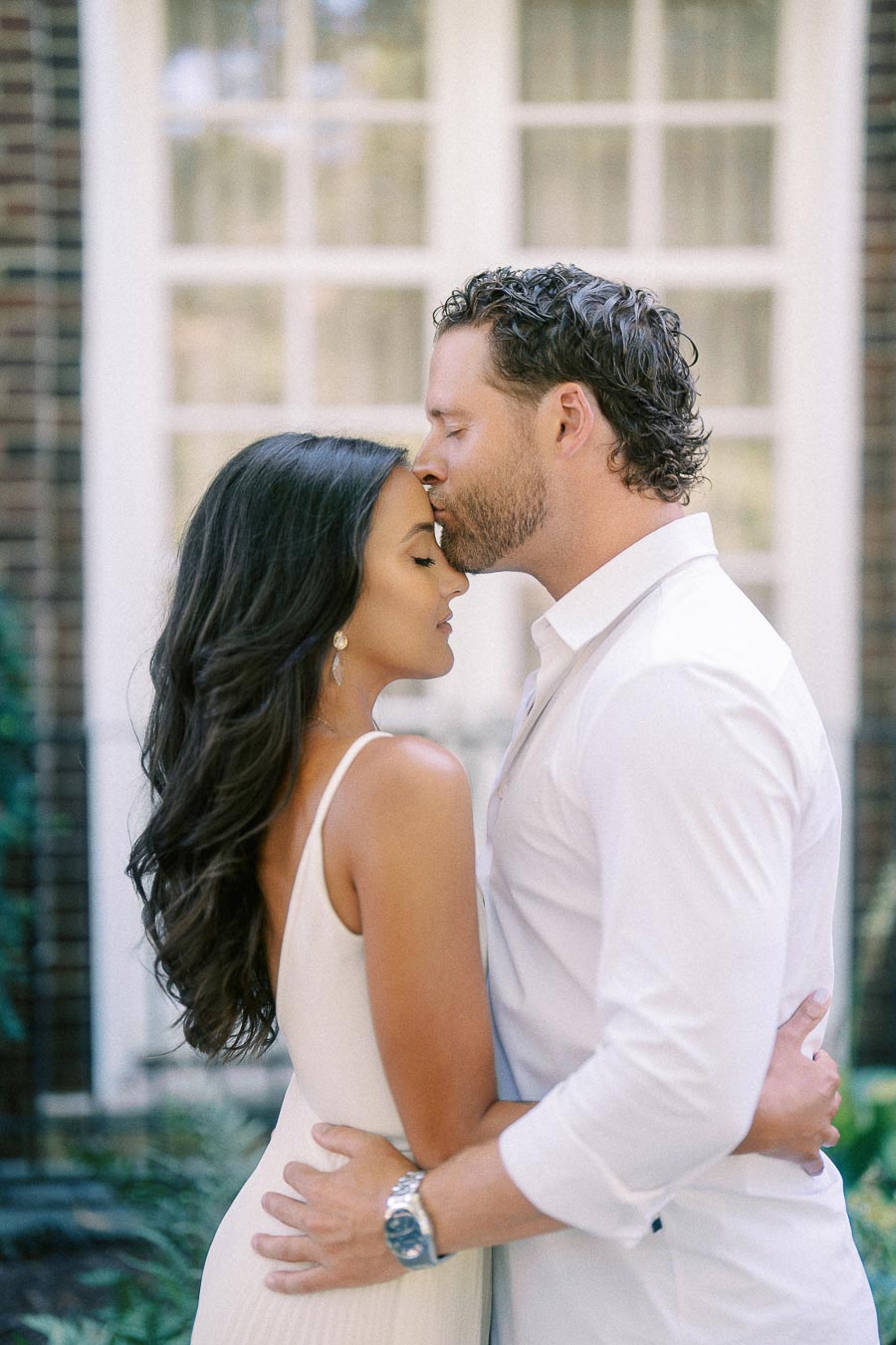 Matt kisses Rachel’s forehead as they embrace beside tall windows in Aurora, Ontario.