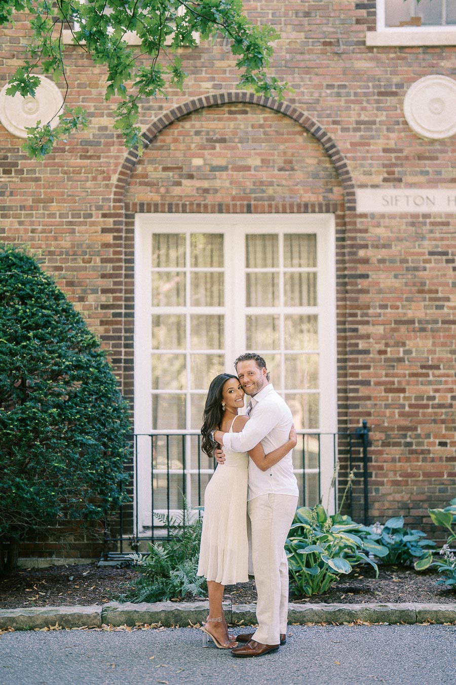 Rachel and Matt embrace in front of a brick building with tall windows and leafy greenery in Aurora.