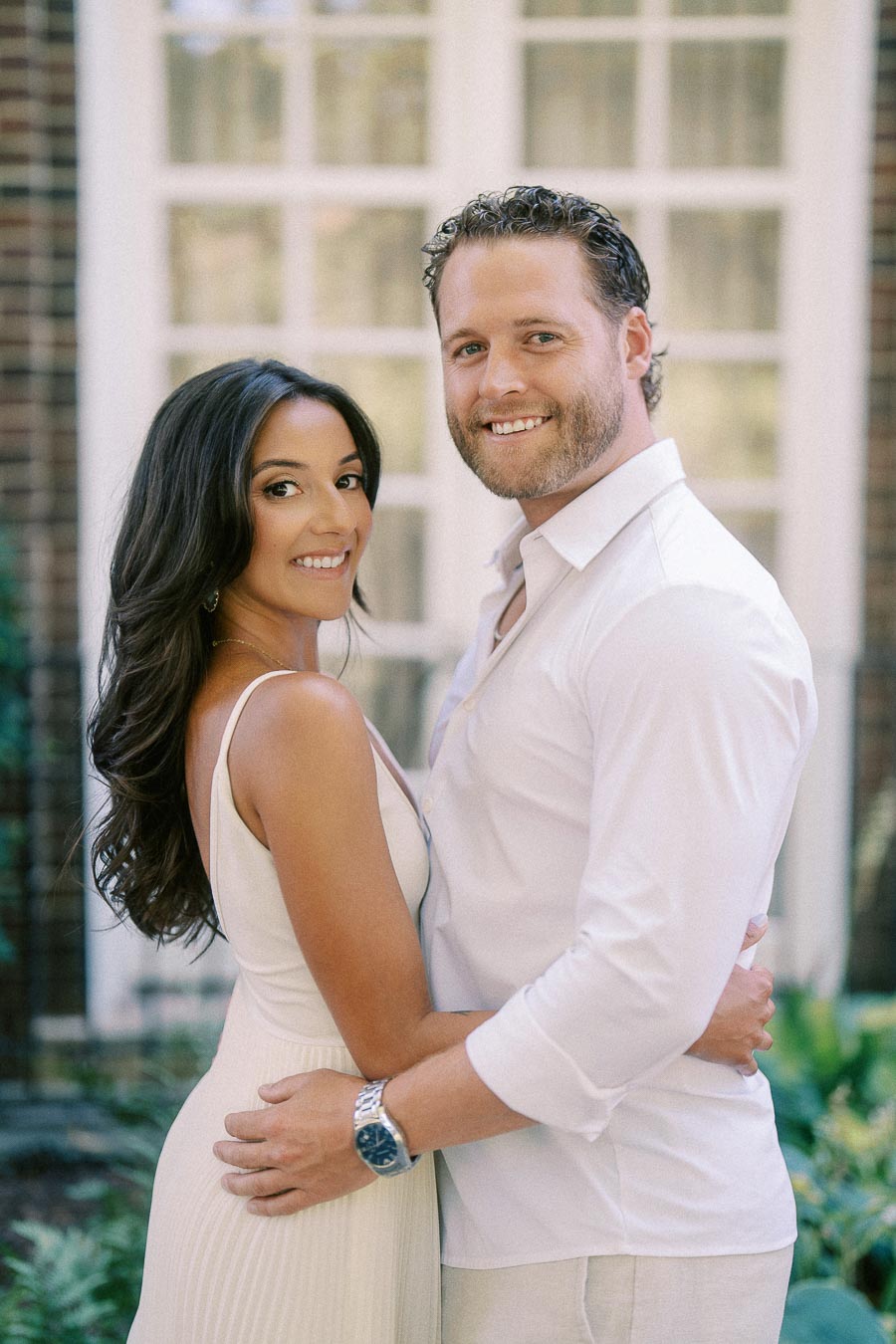 Rachel and Matt smile while embracing in front of tall windows during their engagement session.