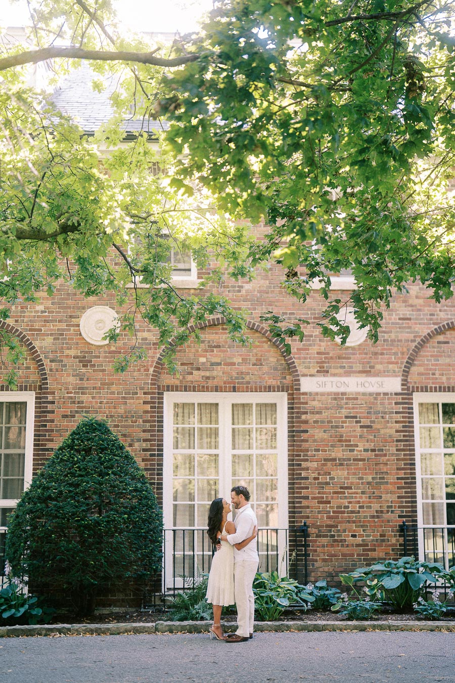 Rachel and Matt embrace beneath leafy trees in front of a brick building with tall windows in Aurora.