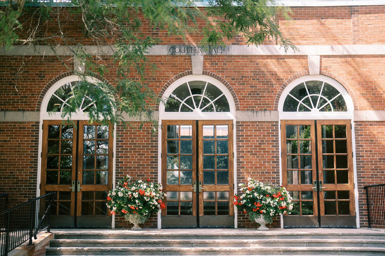 Brick Coulter Hall facade with three arched wooden doors, flower planters, steps, and tree shade.
