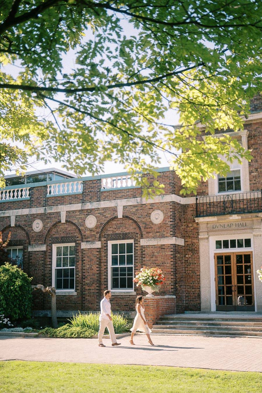 Rachel and Matt walk past Dunlap Hall beneath leafy branches on a sunny campus path.