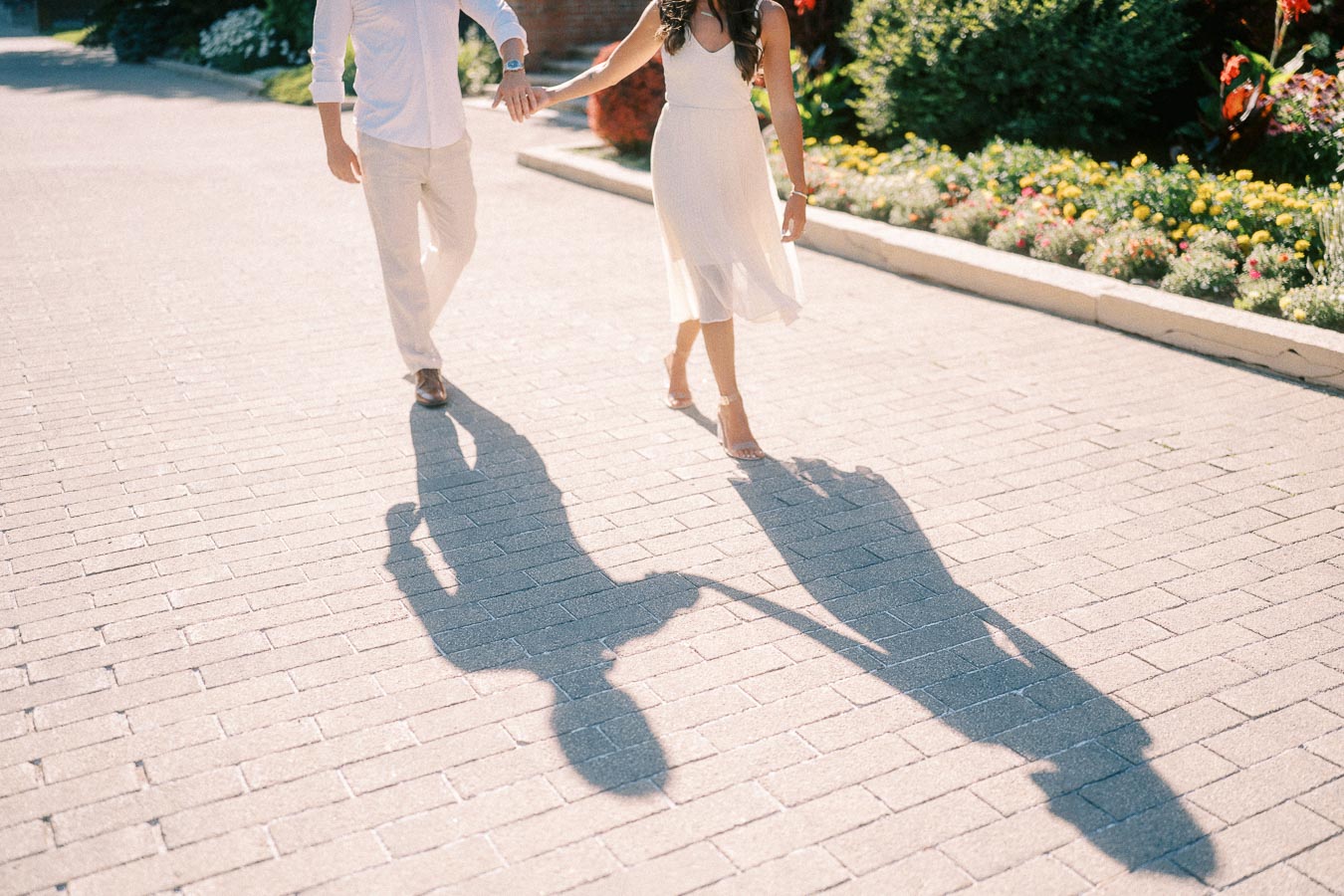 Rachel and Matt walk hand in hand on a sunny brick path, their long shadows stretching across the pavement.