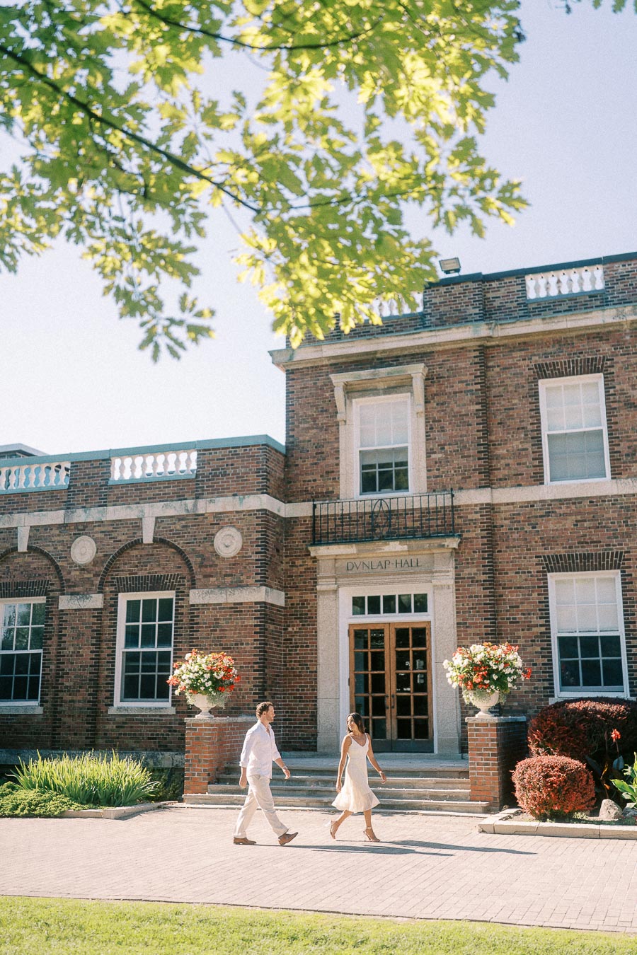 Rachel and Matt walk past the brick entrance of Dunlap Hall beneath sunlit tree branches.