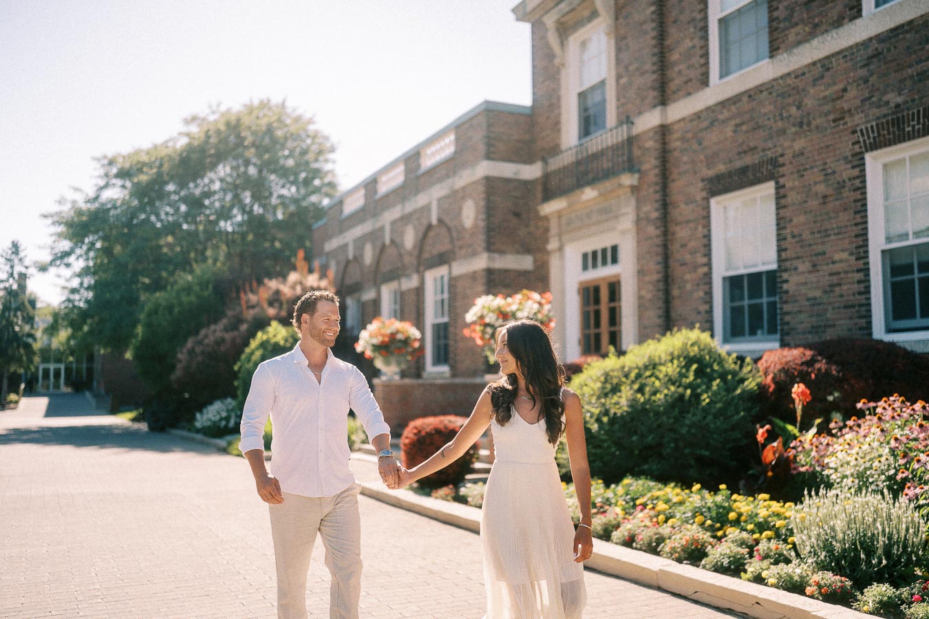 Rachel and Matt walk hand in hand past gardens beside a brick school building in Aurora, Ontario.