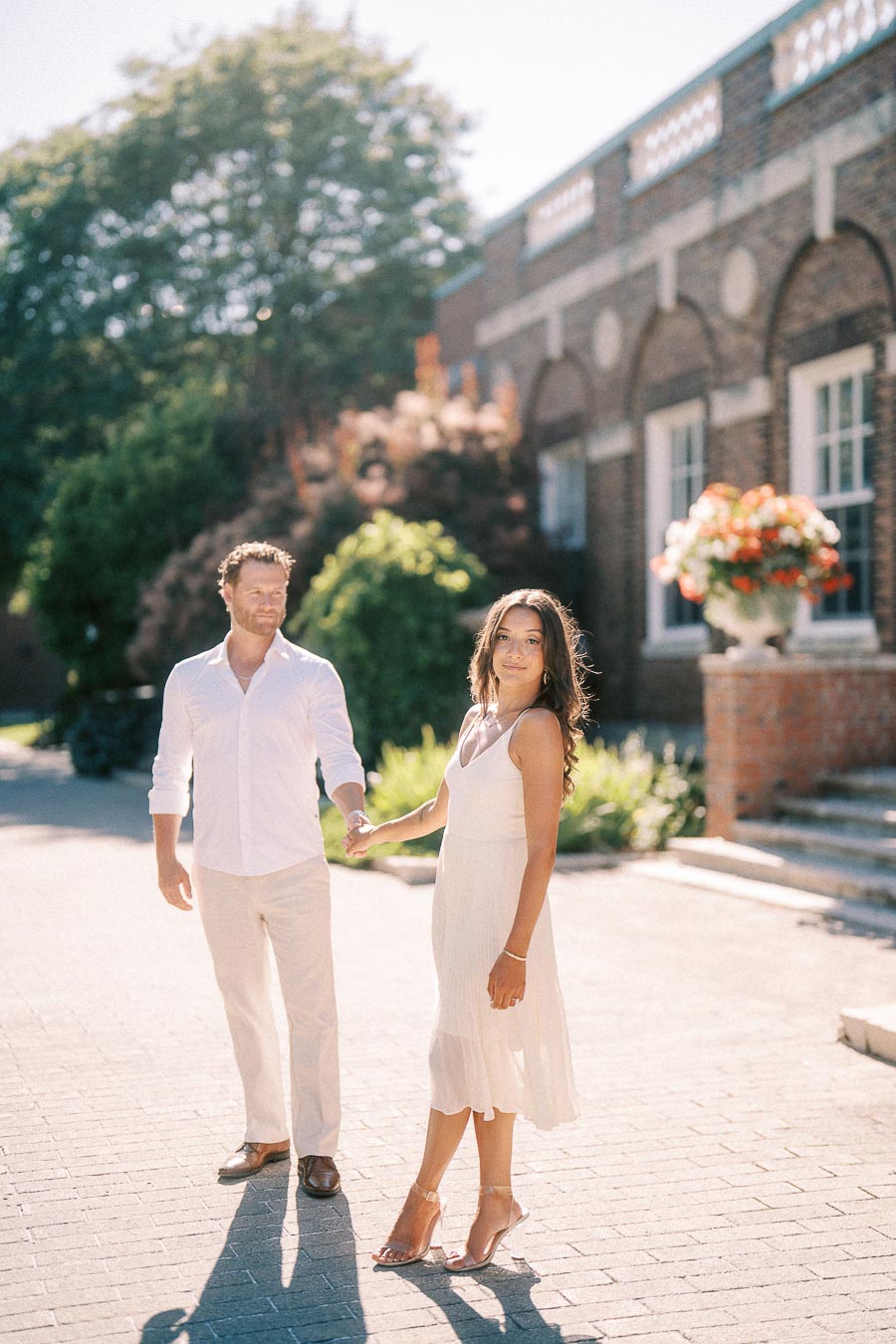 Rachel and Matt hold hands on a sunlit brick walkway beside arched school architecture in Aurora, Ontario.