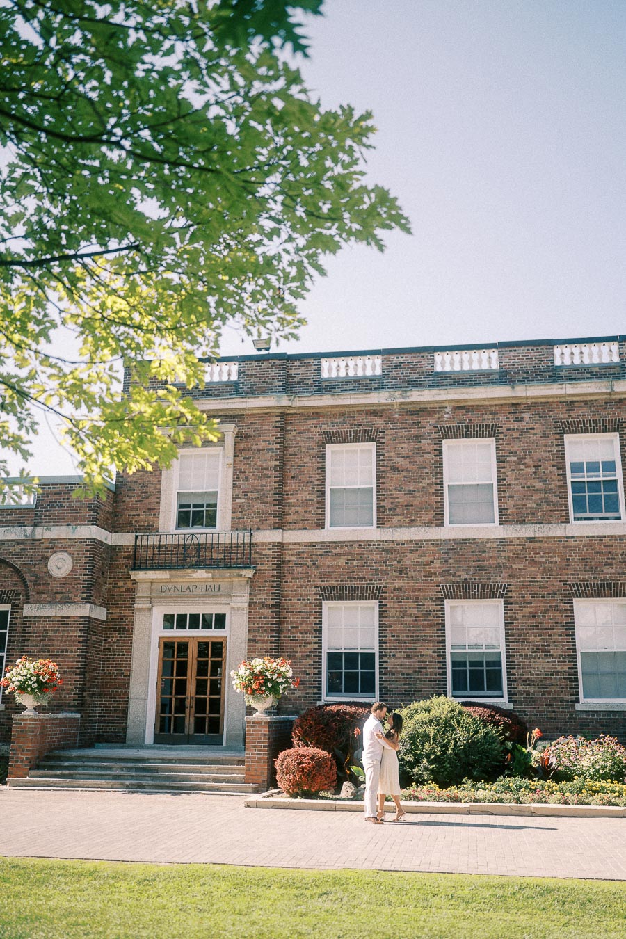Rachel and Matt embrace in front of Dunlap Hall's brick facade, gardens, and leafy trees.