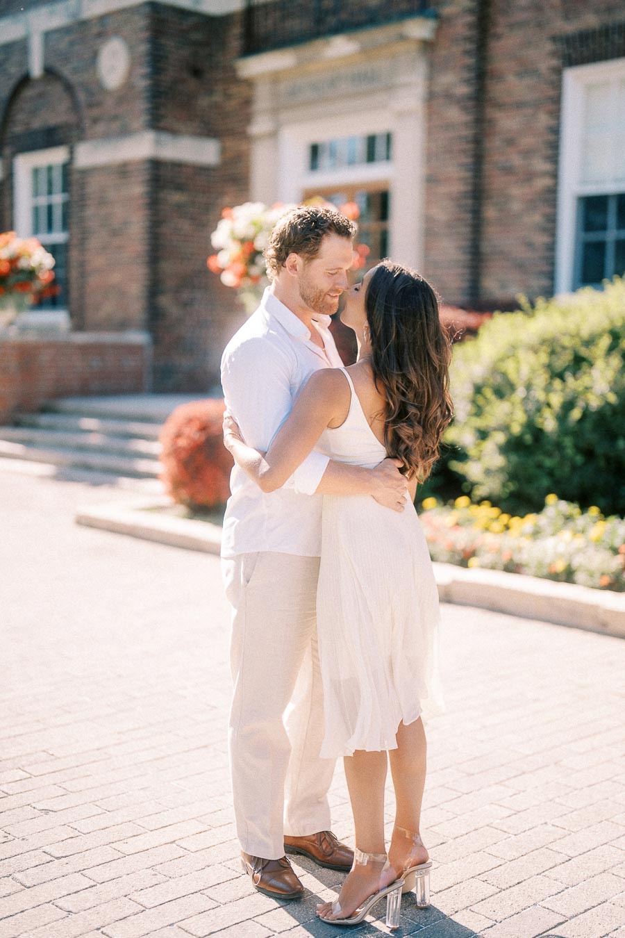 Rachel and Matt embrace closely on a sunlit walkway outside a brick school building in Aurora, Ontario.