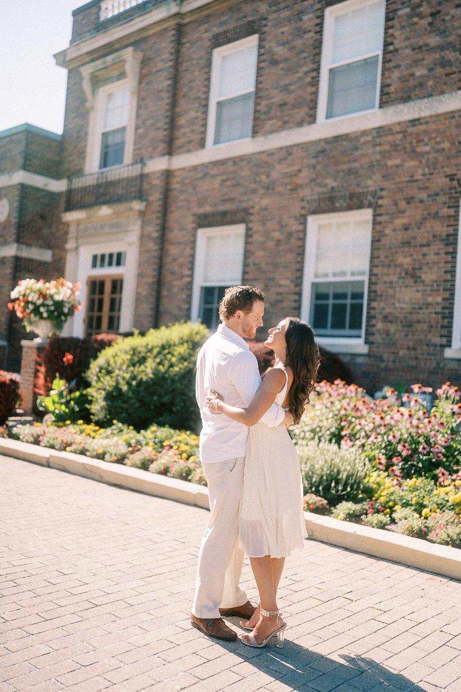 Rachel and Matt embrace on a sunny garden walkway in front of a brick school building in Aurora.
