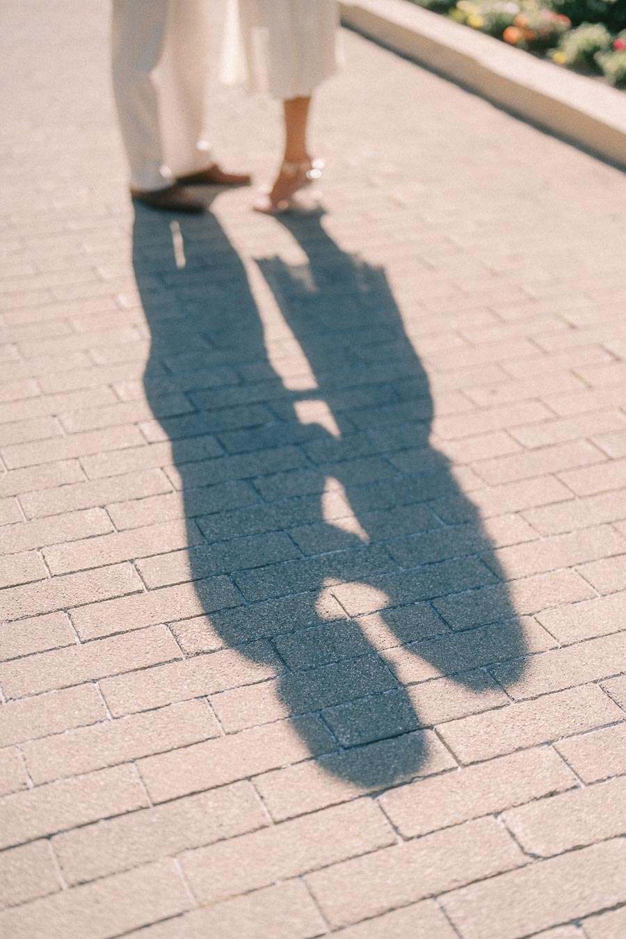 Rachel and Matt stand close on a sunny brick walkway, their shadows stretching across the pavement.