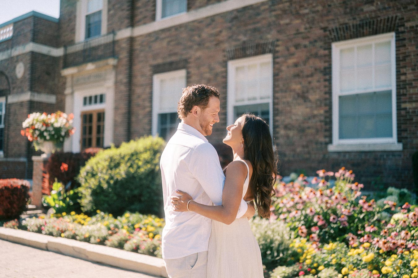 Rachel and Matt smile in an embrace beside bright flower beds and a brick school building in Aurora.