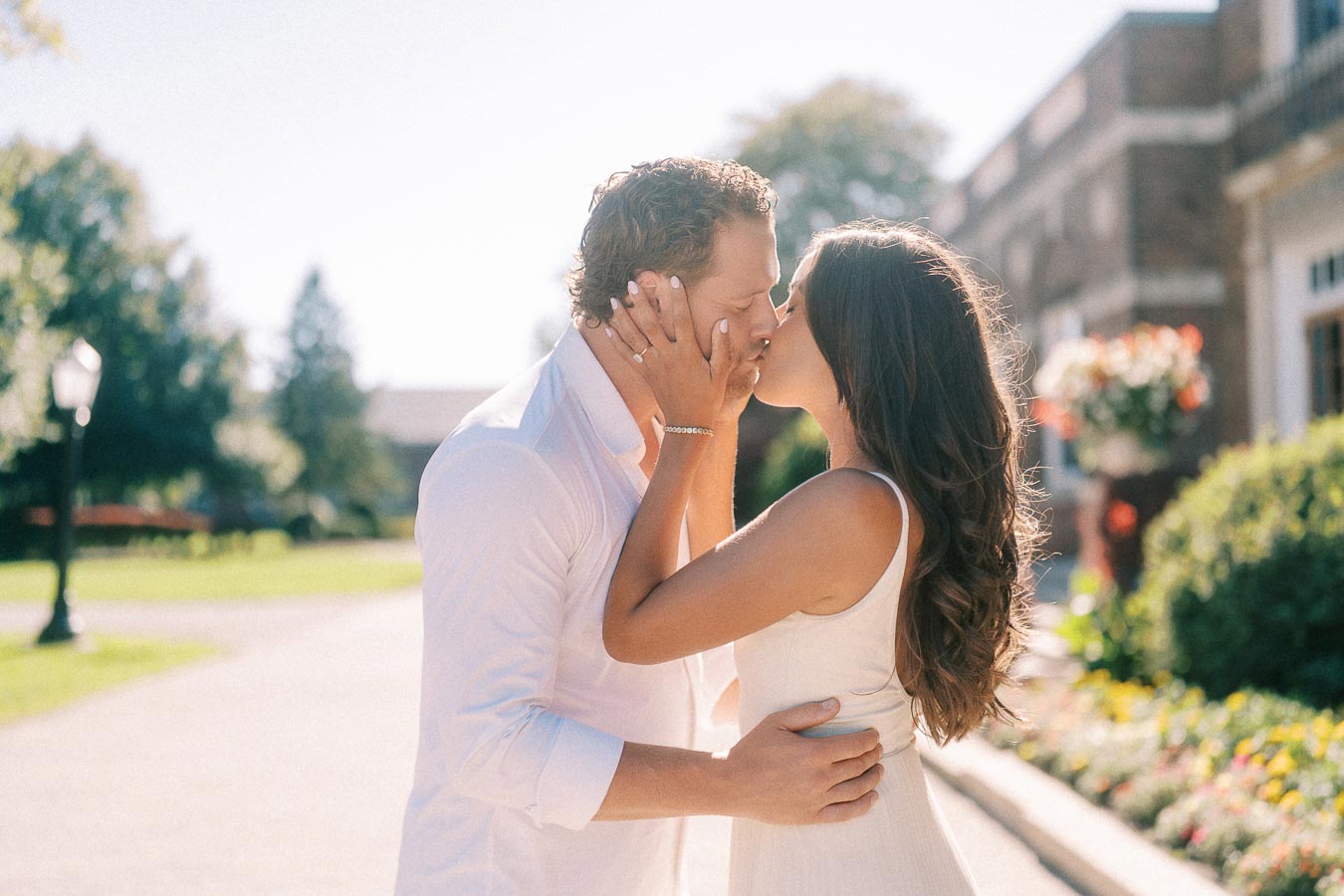 Rachel and Matt kiss on a sunny campus walkway with gardens and a brick building behind them.