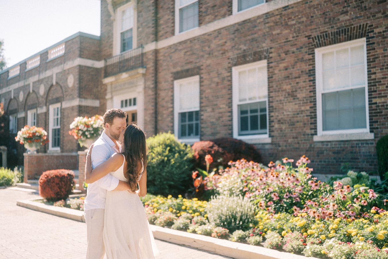 Rachel and Matt embrace beside flower gardens in front of a brick school building in Aurora, Ontario.