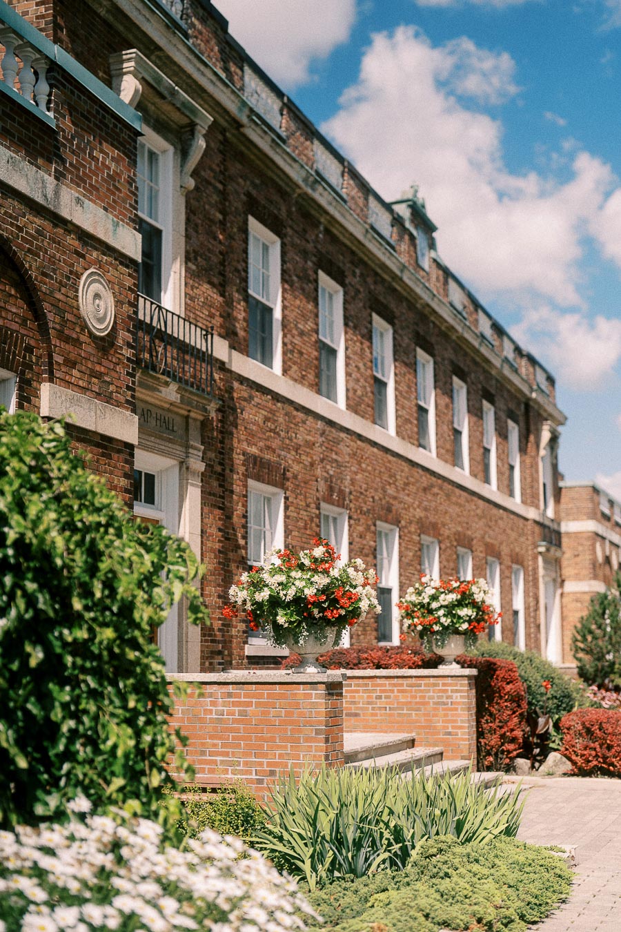 Brick school building with white-trimmed windows, flower planters, and landscaped gardens under a blue sky.
