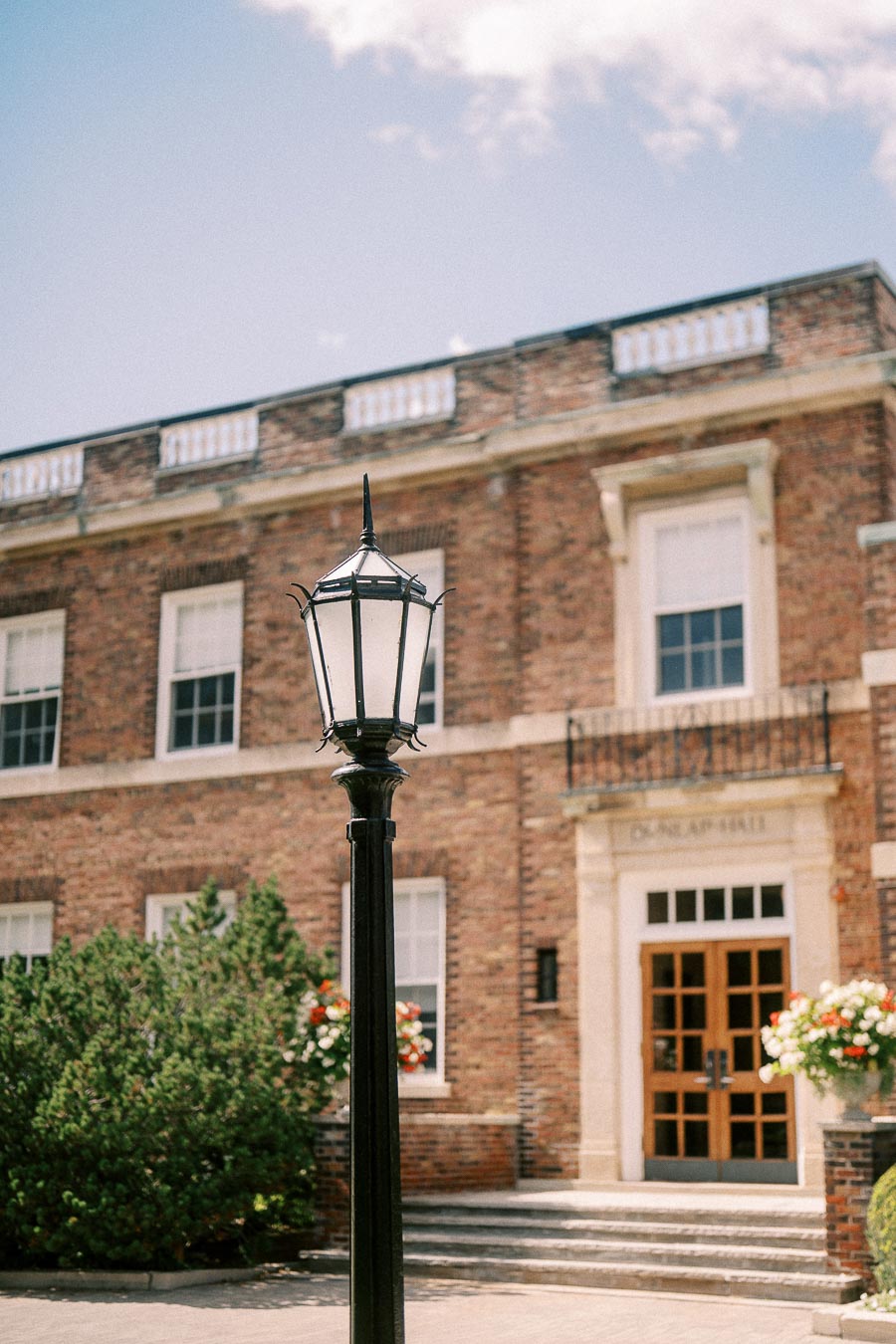 Black lantern post stands before a brick campus building with wooden doors and flower planters.