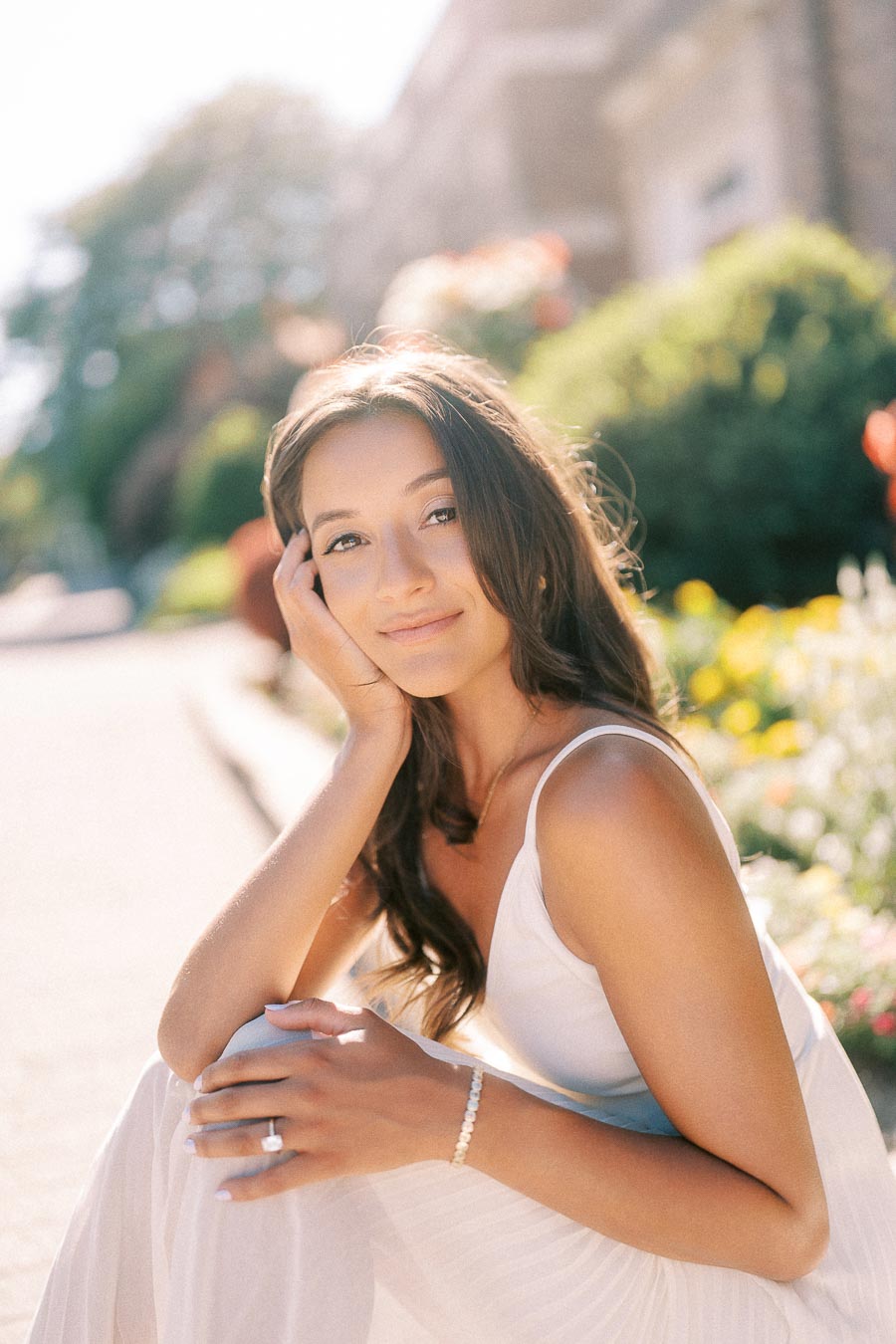 Rachel sits outdoors in a white dress, resting her face in her hand with her engagement ring visible.