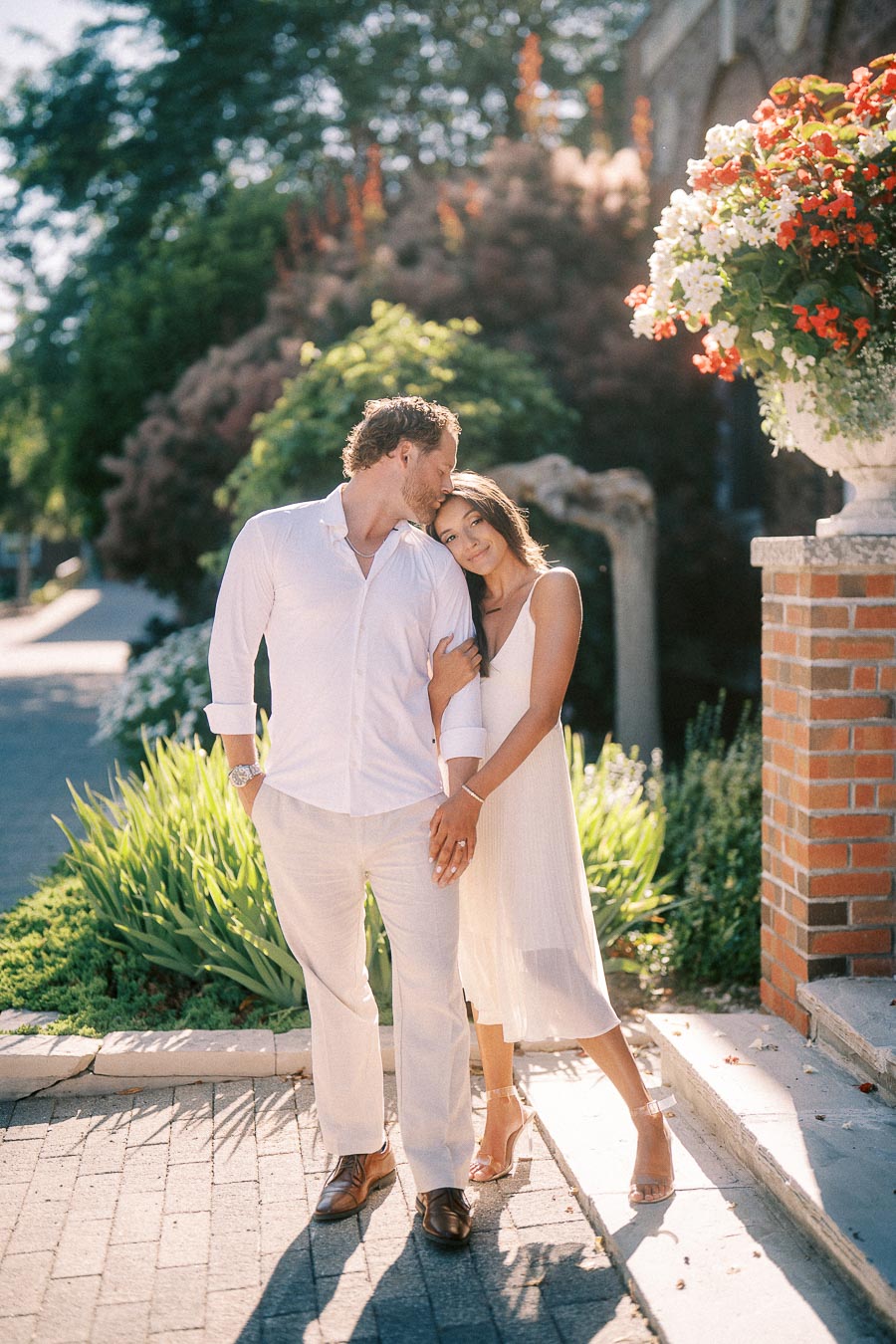 Rachel and Matt stand arm in arm on a sunny garden walkway beside a brick pillar and hanging flowers.