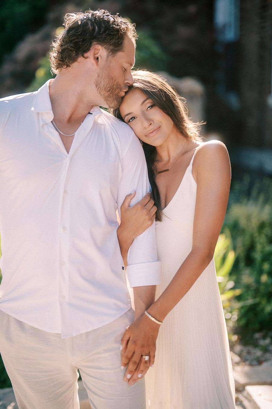 Rachel leans into Matt as he kisses her forehead, both dressed in white outdoors in soft evening light.