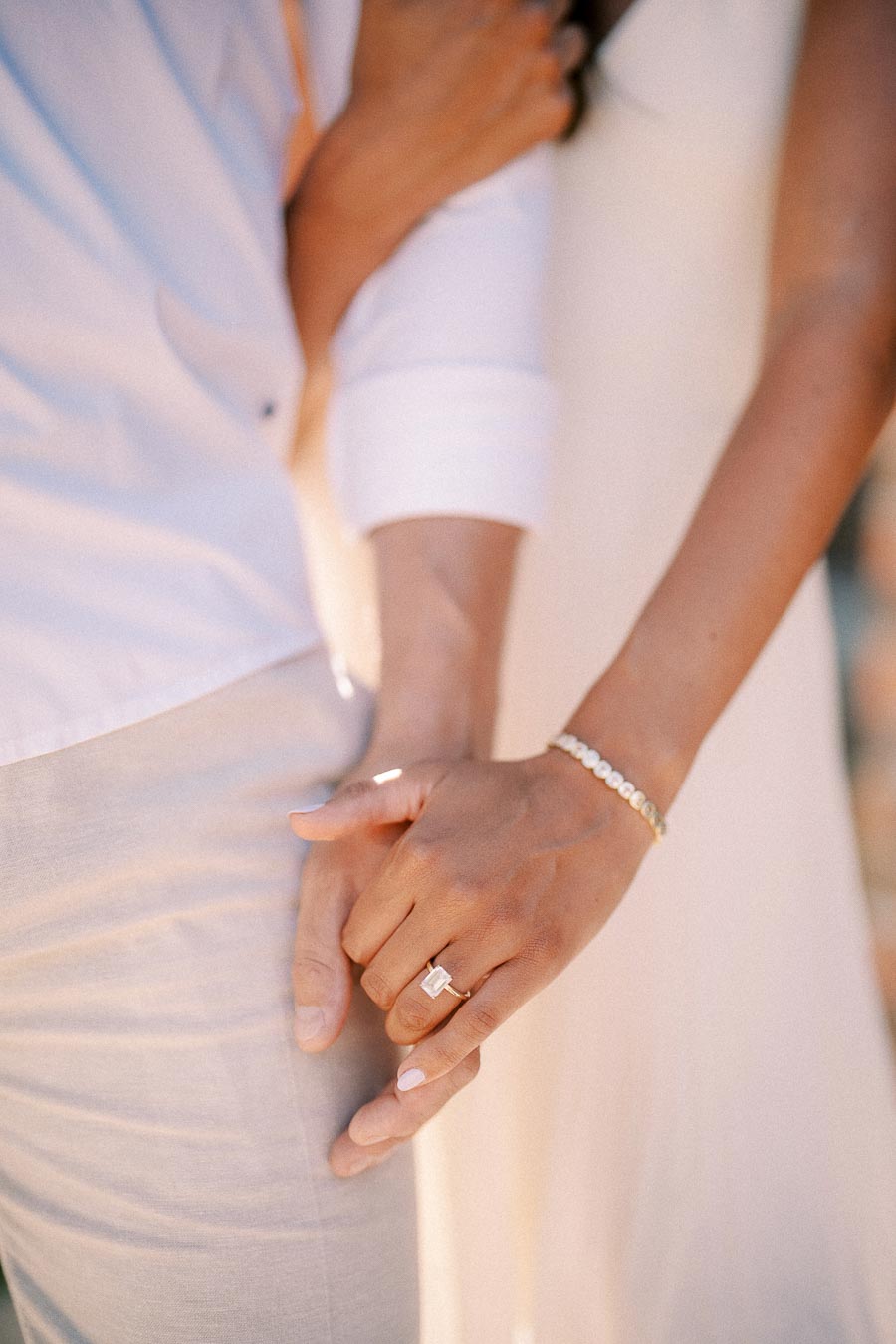 Rachel and Matt hold hands in warm light, with her engagement ring and bracelet in focus.