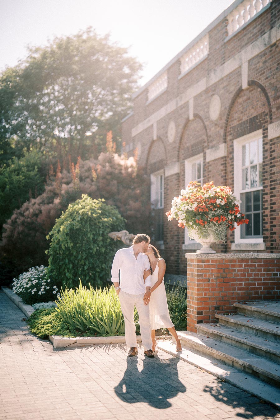 Rachel and Matt kiss beside garden steps and a brick building in warm afternoon light.
