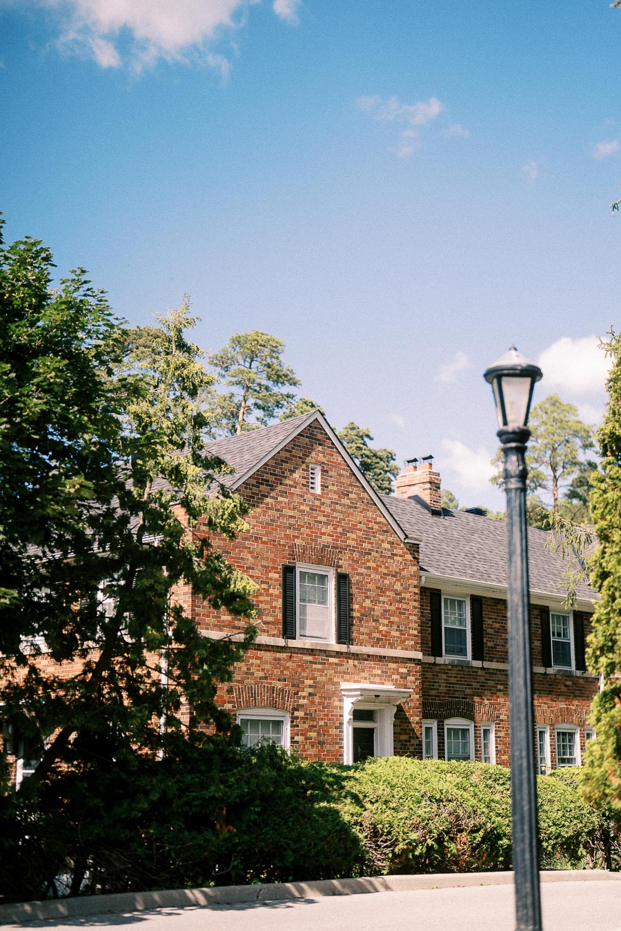Brick campus building with black shutters, hedges, trees, and a lamppost under a blue summer sky.