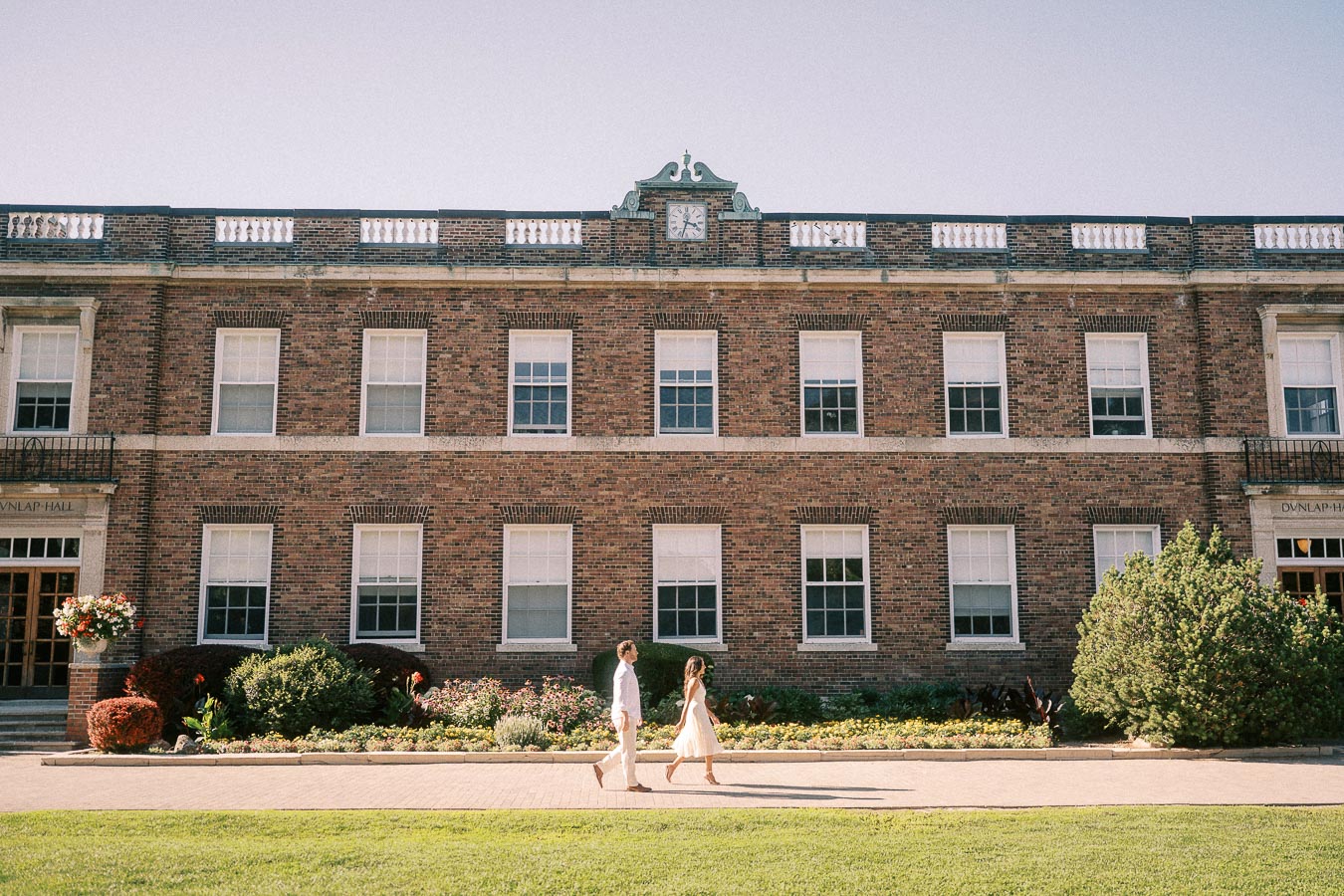 Rachel and Matt walk together in front of a brick school building with tall windows and gardens.