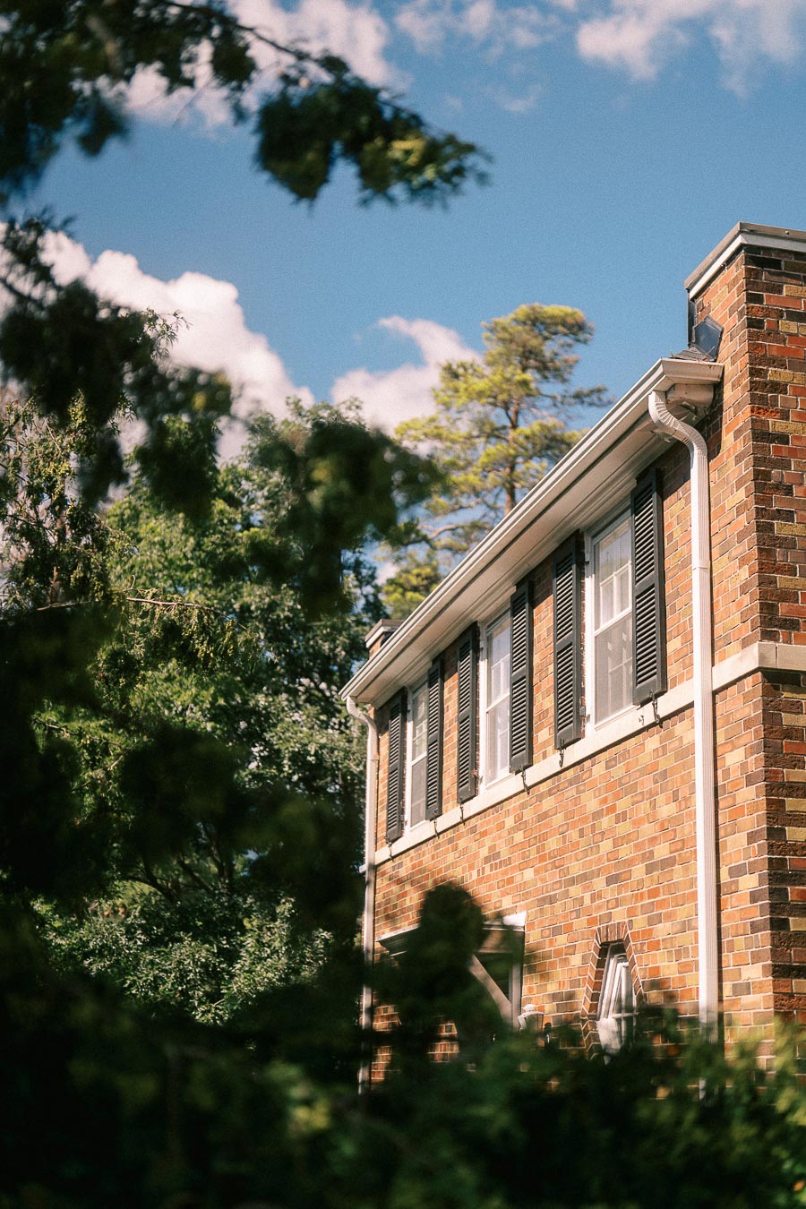 Sunlit brick campus building with shuttered windows seen through trees under a blue sky.