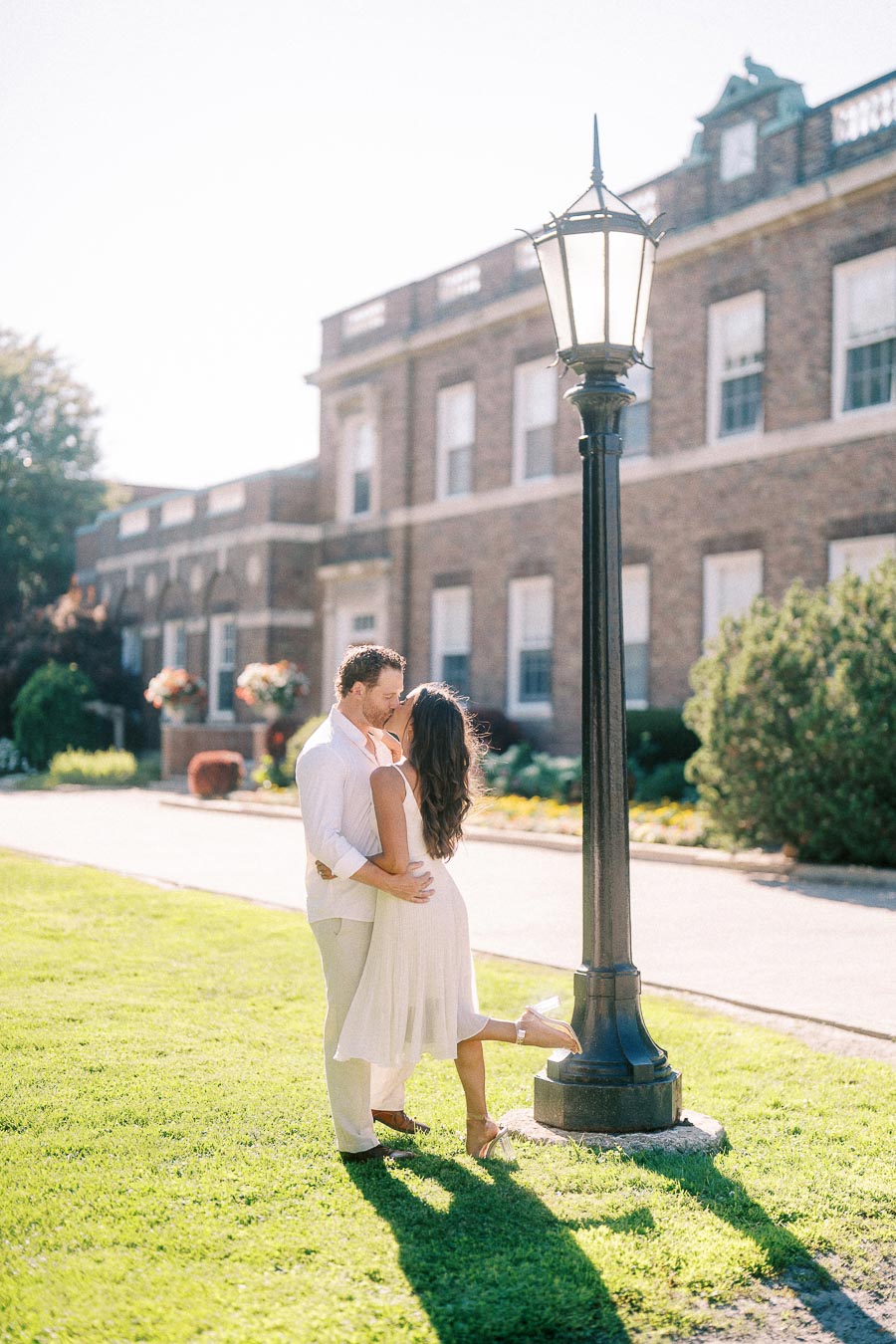 Rachel and Matt embrace beside a lamp post on a sunny lawn in front of a brick campus building.