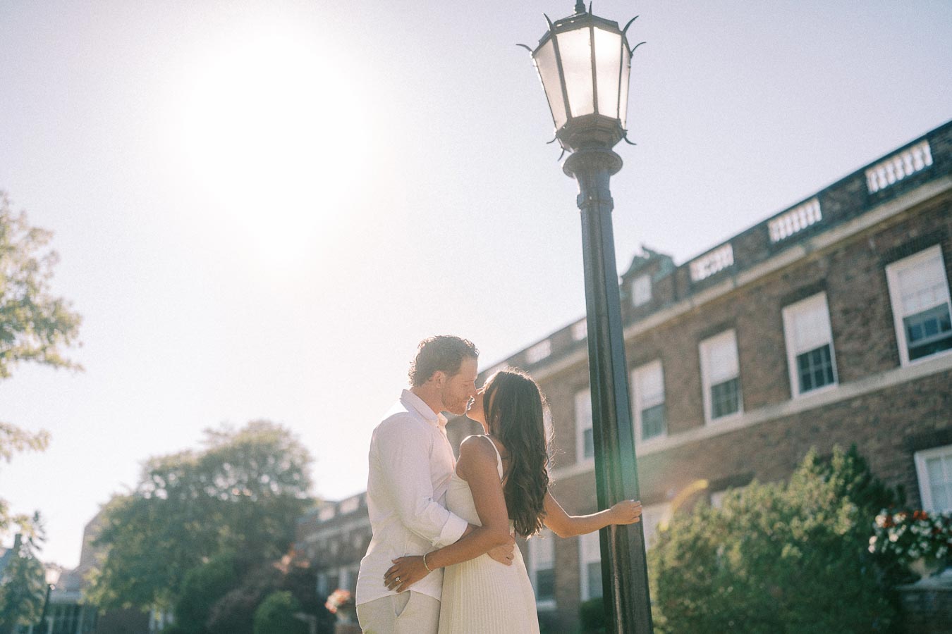 Rachel and Matt kiss beside a tall lamppost in front of a brick campus building in bright sunlight.