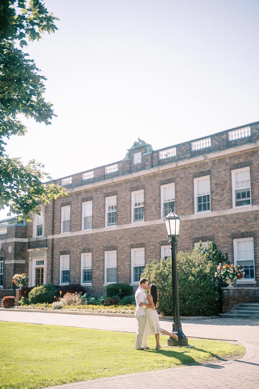 Rachel and Matt embrace beside a lamp post on a sunny lawn in front of a brick school building.