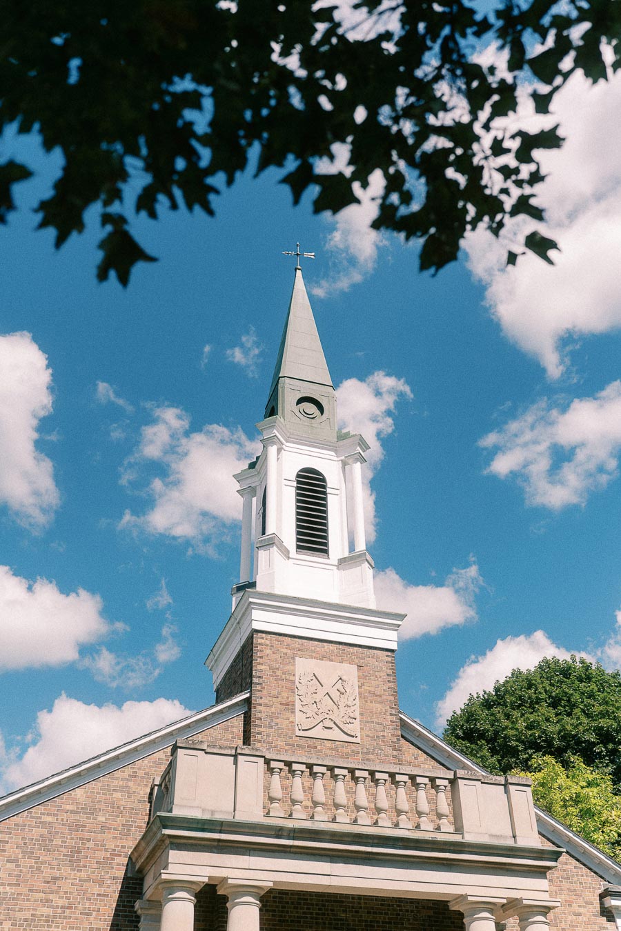White chapel steeple rises above a brick building, framed by tree leaves against a blue sky.
