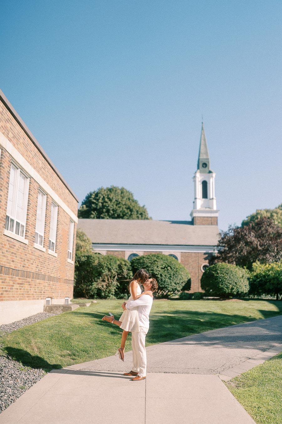 Matt lifts Rachel on a sunny campus walkway with brick buildings, lawns, and a chapel steeple behind them.