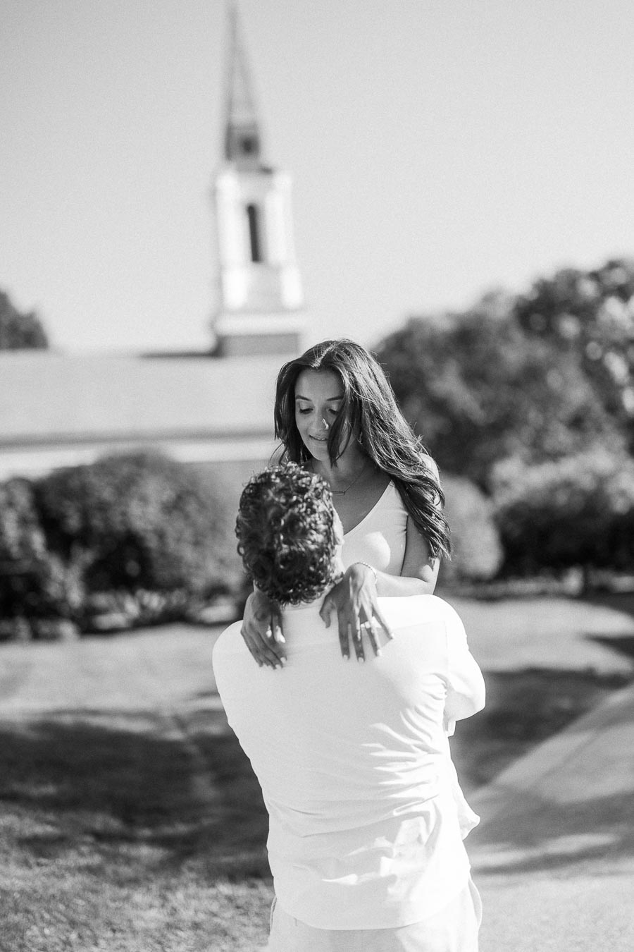 Matt lifts Rachel in his arms outdoors, with a blurred chapel steeple and lawns behind them.