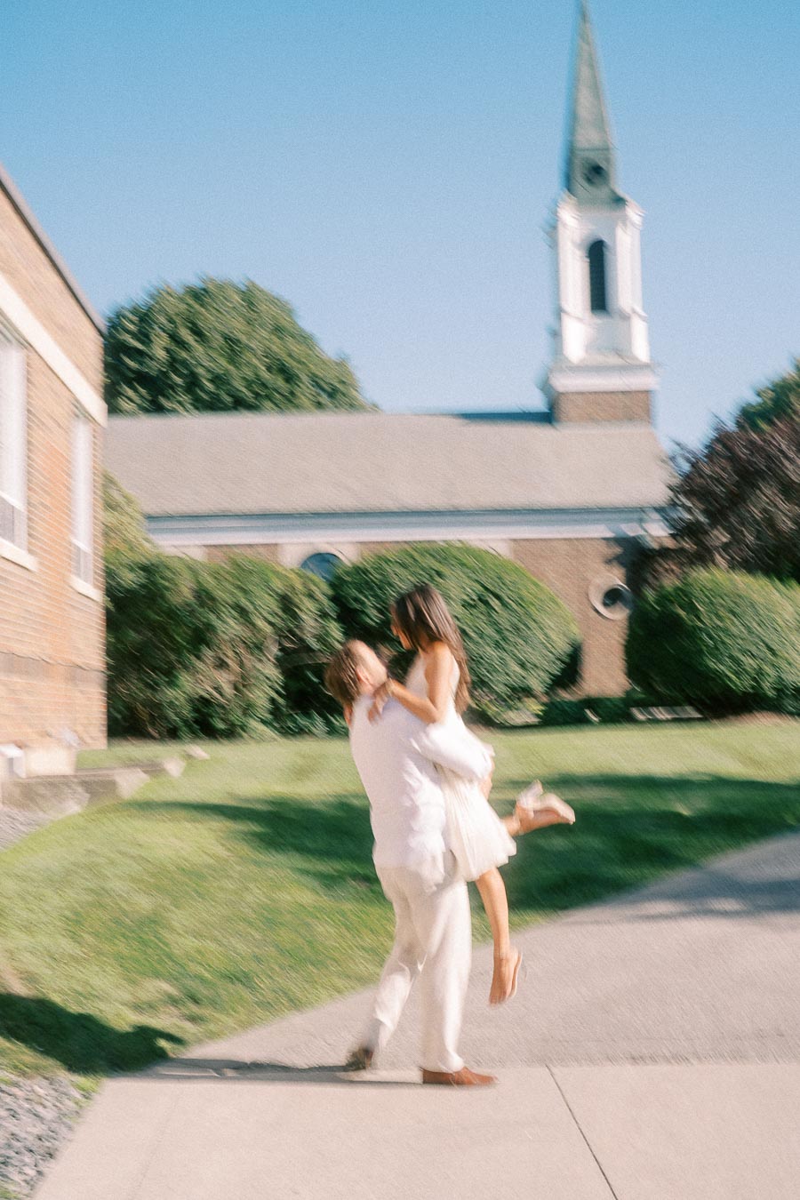 Rachel and Matt embrace as he lifts her on a sunlit campus walkway near a chapel in Aurora.