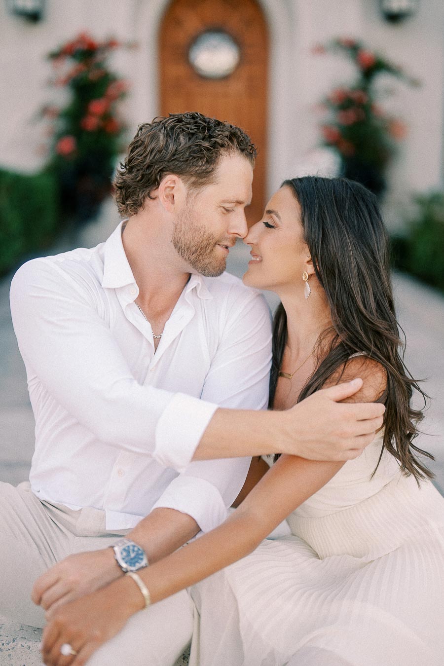 Rachel and Matt sit close together, smiling nose to nose in front of an arched doorway and flowers.