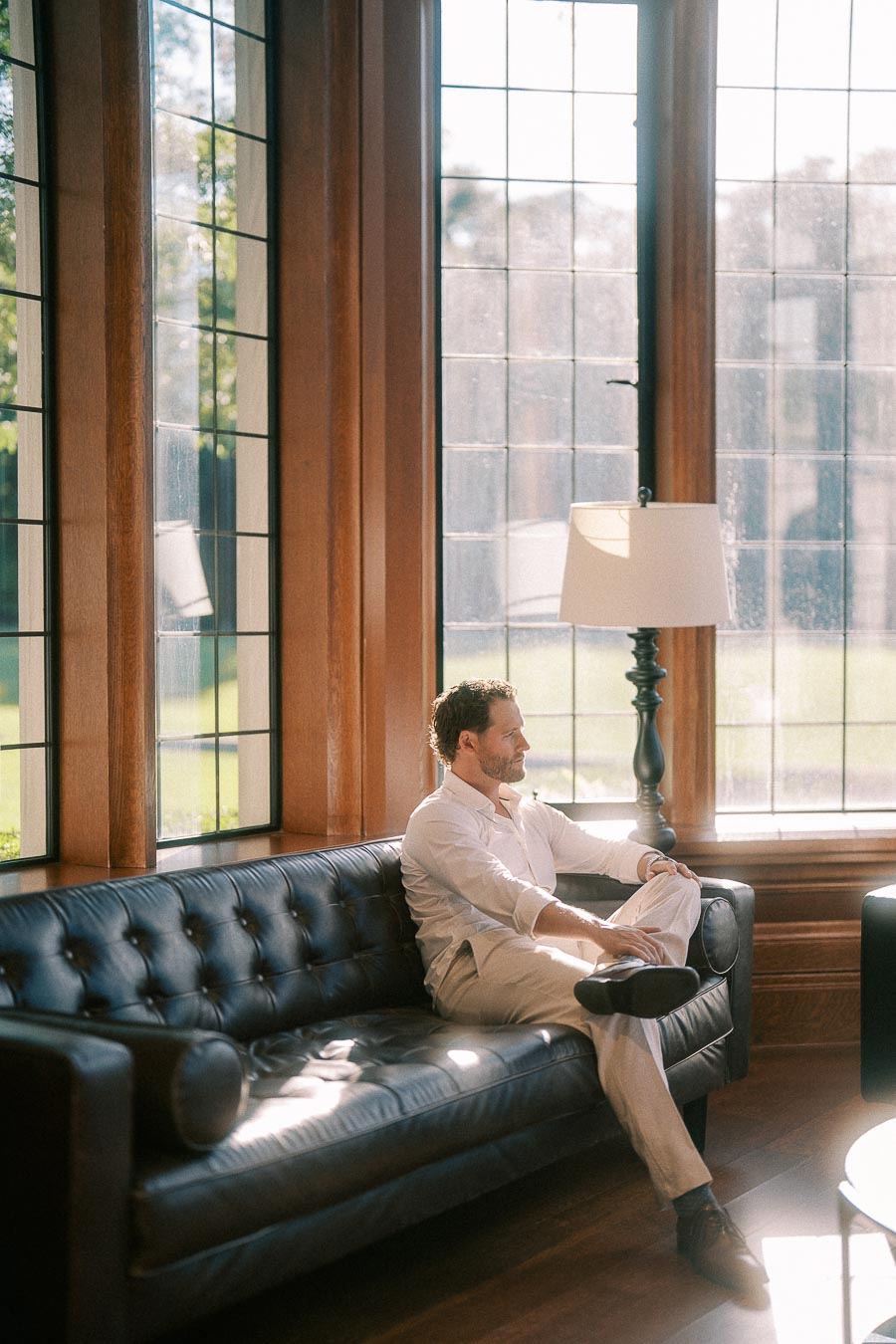 Matt sits on a black leather sofa beside tall leaded windows in a sunlit wood-paneled room.
