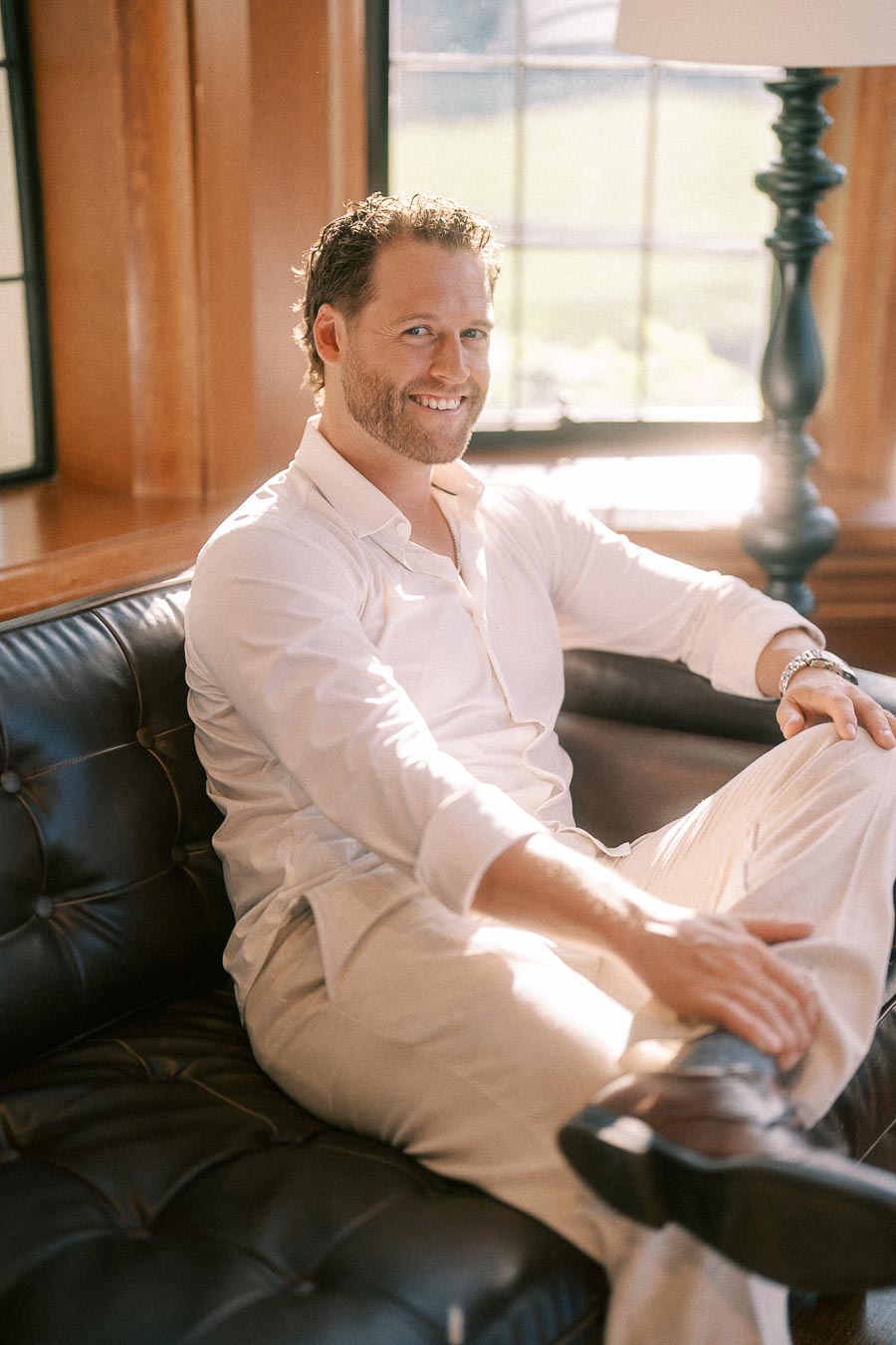 Matt sits smiling on a black leather couch in a sunlit wood-paneled room with tall windows.