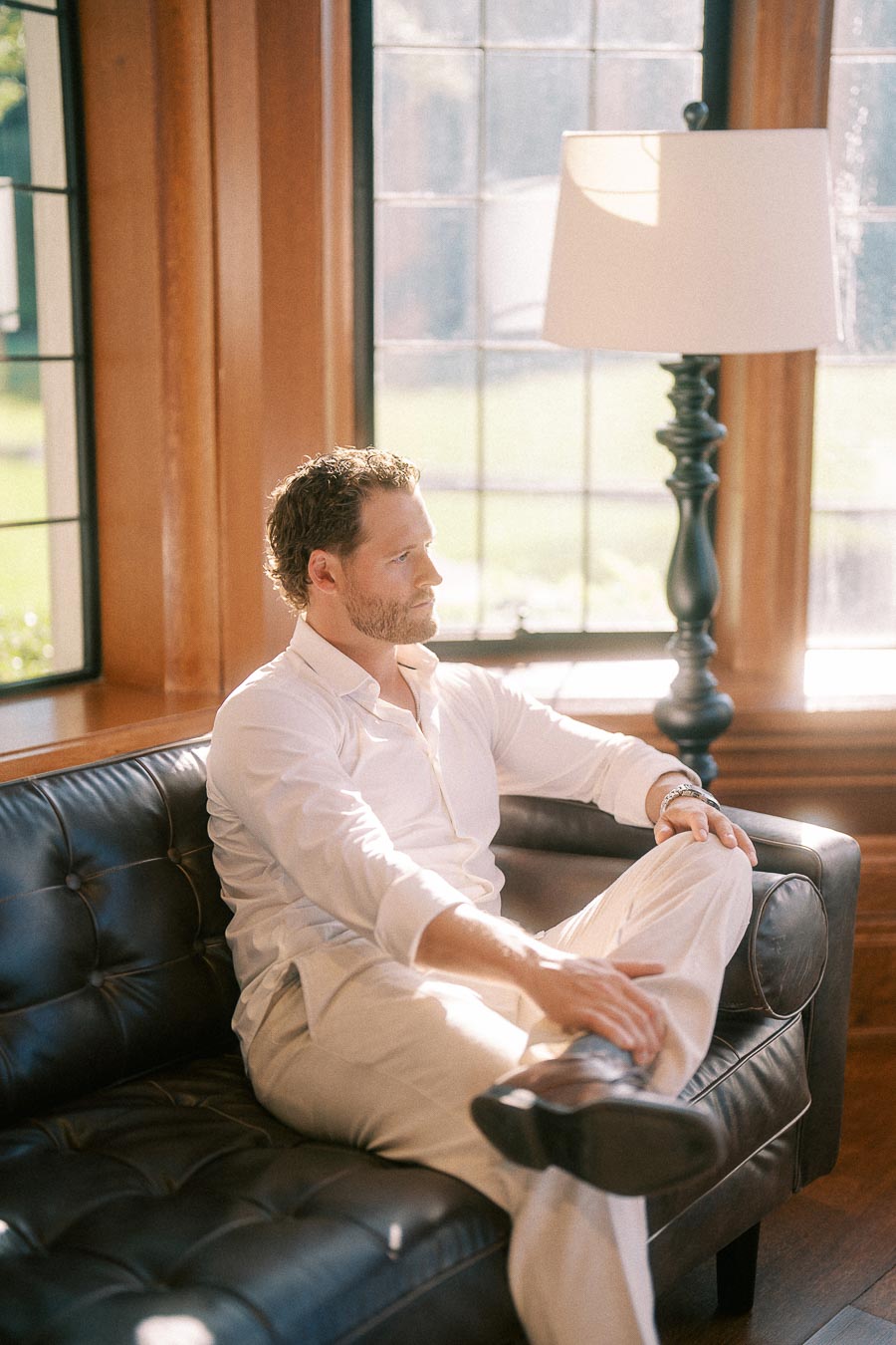 Matt sits on a black leather sofa in a sunlit wood-paneled room with tall windows and a floor lamp.