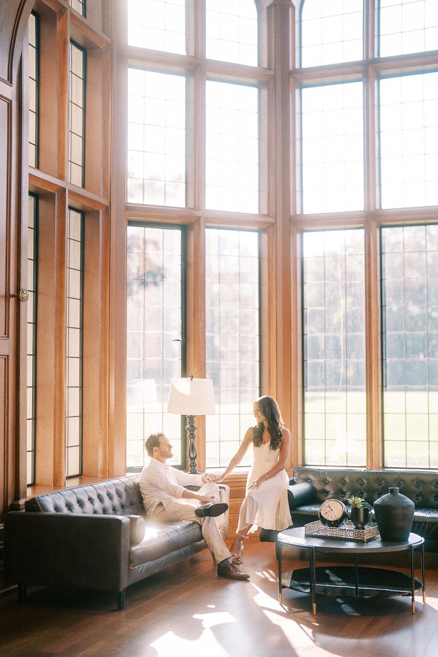 Rachel and Matt hold hands on leather sofas in a sunlit room with tall wood-framed windows.