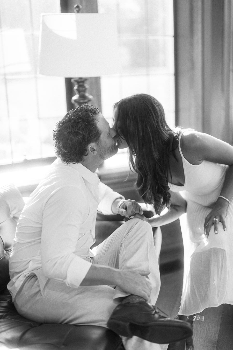 Rachel and Matt kiss beside a bright window while sitting together on a leather sofa indoors.