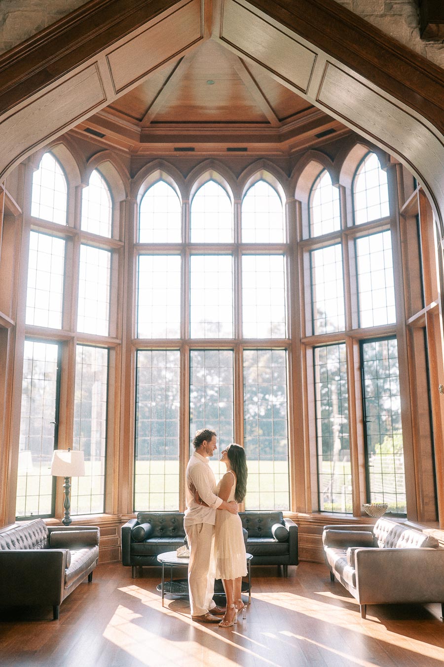 Rachel and Matt stand together in a sunlit wood-paneled room with tall arched windows and sofas.