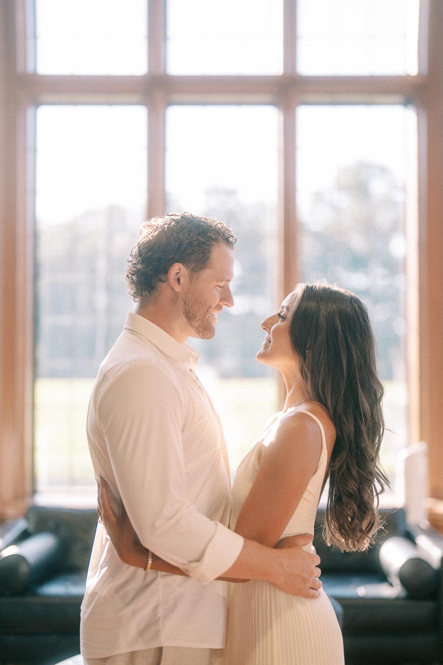 Rachel and Matt smile at each other while embracing indoors in front of tall sunlit windows.