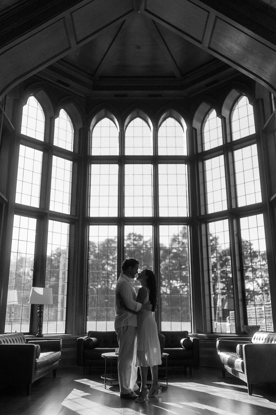 Rachel and Matt embrace in silhouette before tall arched windows in a wood-paneled room.