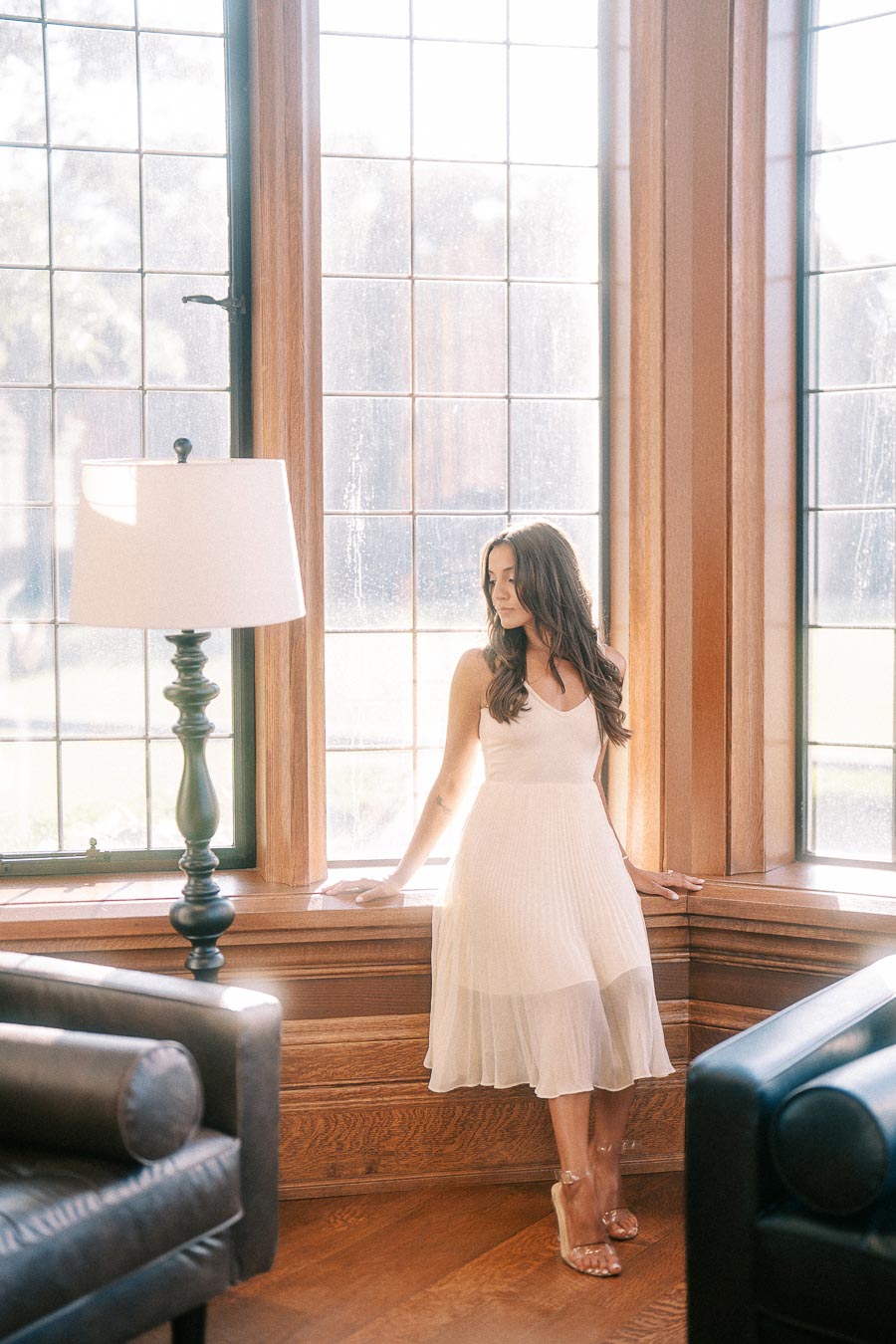 Rachel sits by tall wood-framed windows in a white dress, with leather chairs and a floor lamp nearby.