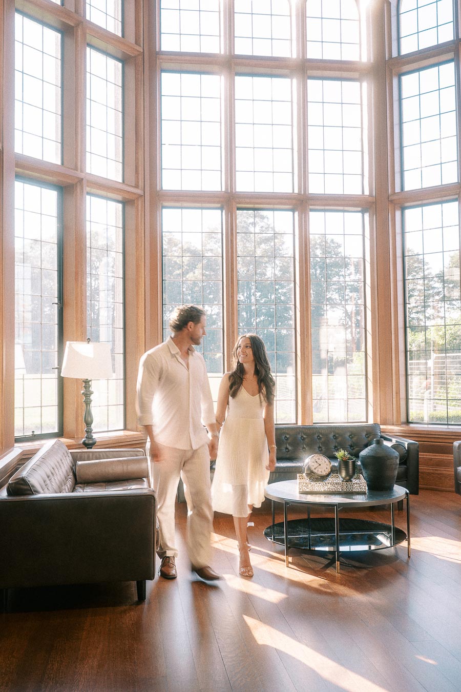Rachel and Matt hold hands in a sunlit lounge with tall grid windows, sofas, and wood floors.