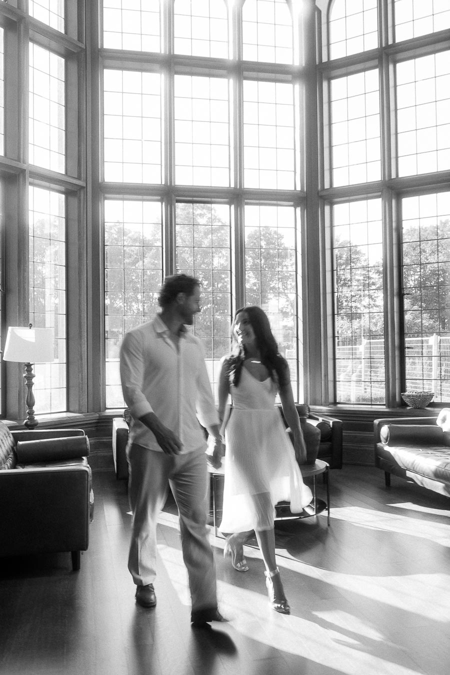 Rachel and Matt walk hand in hand through a bright lounge with tall grid windows and sunlit floors.