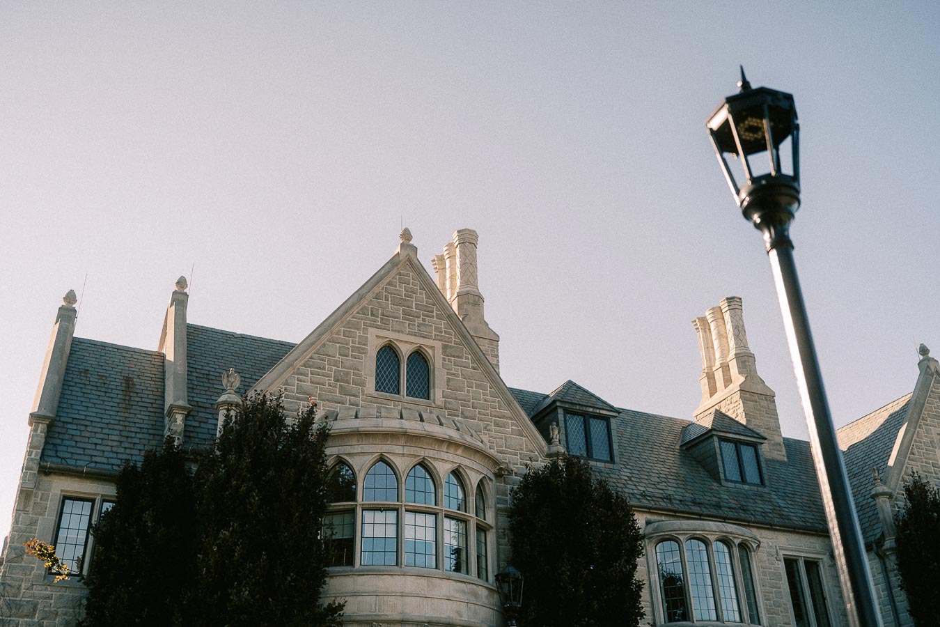 Stone collegiate building with arched windows, steep slate roof, chimneys, and a lamppost at dusk.