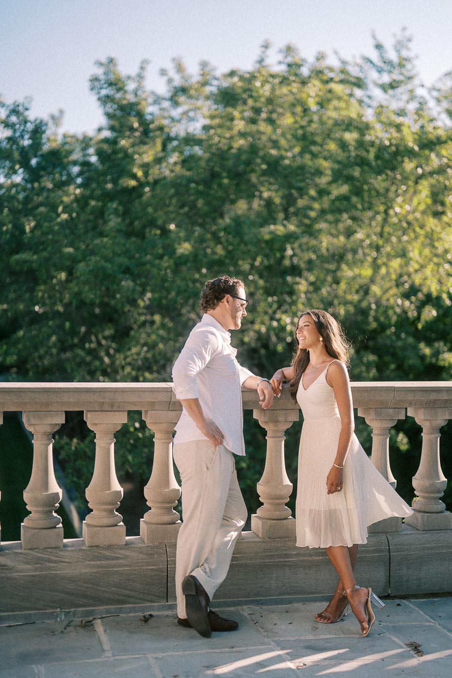 Rachel and Matt smile together beside a stone balustrade with sunlit trees behind them.